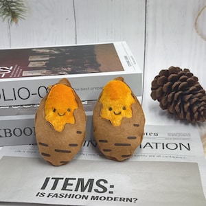 May include: Two plush toys shaped like sweet potatoes, each with a smiling face and orange filling. The toys are brown with a textured design and sit on a newspaper with the text "ITEMS: IS FASHION MODERN?"
