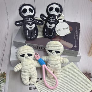 Puede incluir: Cuatro llaveros de peluche con temática de Halloween. Dos son figuras de esqueletos negros con detalles de huesos blancos. Los otros dos son figuras de momias envueltas en tela de color crema. Un llavero tiene una etiqueta que dice "your text".
