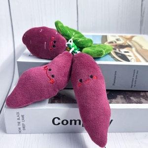 May include: Plush sweet potato toys in shades of purple and green. The sweet potatoes have embroidered smiling faces and are attached to green plush leaves. The toys are resting on top of books.