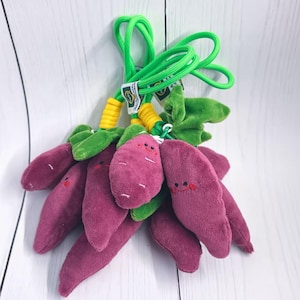 May include: A cluster of plush purple sweet potatoes with green leaves and smiling faces, attached to a green rope. The sweet potatoes have embroidered details and are designed as a toy or decorative item.