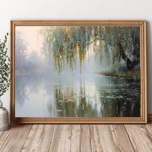 May include: A framed landscape painting depicts a serene lake scene with a weeping willow tree. The artwork features soft, muted colors, with the tree's foliage and reflections in the water. The frame is made of light-colored wood.