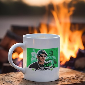 Puede incluir: Taza de cerámica blanca con un rectángulo verde con un gráfico de un hombre con gafas de sol y un sombrero. La taza tiene la palabra "FEID" y la frase "Que chimba so" impresa. La taza está sobre una superficie de madera.