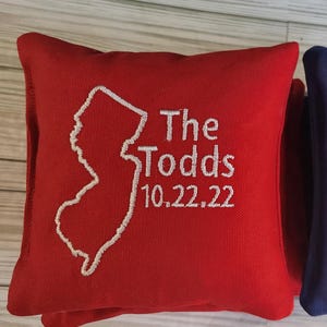 May include: Two square bean bags, one red and one blue, featuring white embroidered outlines of the state of New Jersey. Each bag displays the text The Todds and the date 10.22.22 in white embroidery on the front surface of the fabric.
