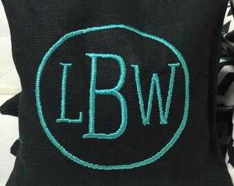 Op de afbeelding: Een vierkante zwarte stoffen zitzak voor werpspellen met een geborduurde blauwgroene cirkelomtrek rondom de letters LBW in bijpassende blauwgroene draad. De stof heeft een zichtbare geweven textuur en het borduurwerk is in het midden geplaatst.