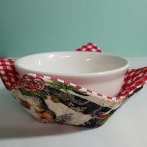 Op de afbeelding: Een witte keramische kom rust in een stoffen komhouder. De houder heeft een rood-wit ruitpatroon op de randen en een bloemen- en dierenprint aan de binnenkant. De houder is ontworpen om de kom vast te houden.