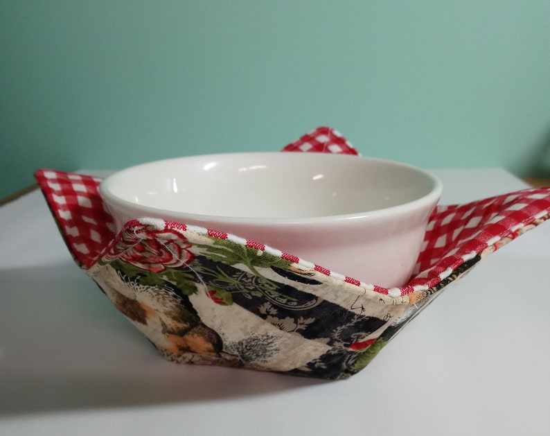 Op de afbeelding: Een witte keramische kom rust in een stoffen komhouder. De houder heeft een rood-wit ruitpatroon op de randen en een bloemen- en dierenprint aan de binnenkant. De houder is ontworpen om de kom vast te houden.