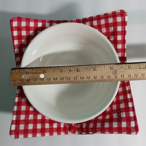 Op de afbeelding: Een witte keramische kom op een rood-wit geruite komhouder. Een houten liniaal meet de diameter van de kom, ongeveer 15 cm. De komhouder is gemaakt van stof.