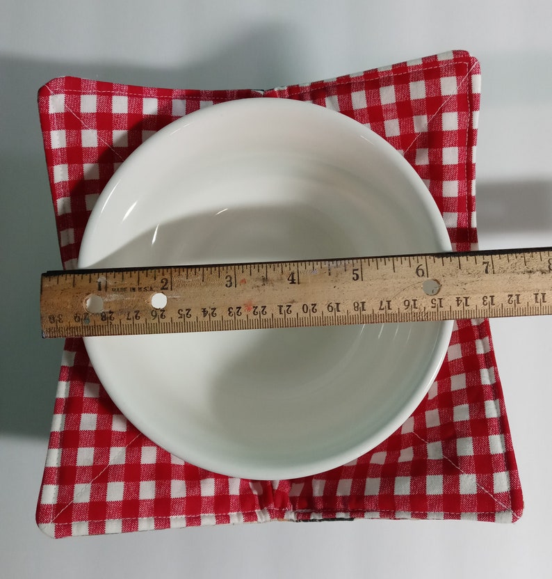 Op de afbeelding: Een witte keramische kom op een rood-wit geruite komhouder. Een houten liniaal meet de diameter van de kom, ongeveer 15 cm. De komhouder is gemaakt van stof.