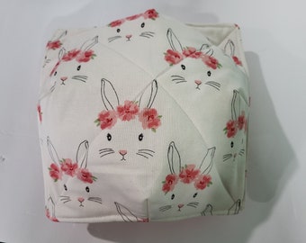 May include: A white fabric container with a repeating pattern of bunny faces wearing floral crowns. The bunnies have black whiskers and eyes, and pink noses. The container is quilted with a diamond pattern.
