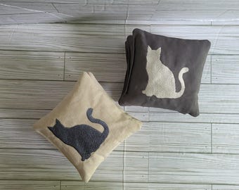 Op de afbeelding: Twee vierkante kussens met silhouetten van katten. Het ene kussen is beige met een donkergrijze kat-applicatie, terwijl het andere donkergrijs is met een beige kat-applicatie. Beide kussens hebben een zacht, getextureerd oppervlak.
