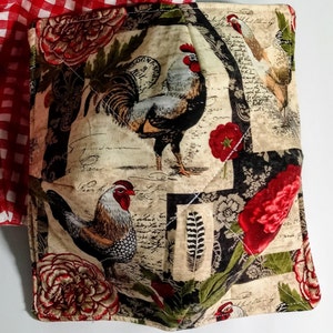 Op de afbeelding: Een gewatteerde stoffen komhoes met een haan- en bloemenontwerp. De stof heeft hanen, rode bloemen en tekst op een cr&egrave;mekleurige achtergrond. Een rood-wit geruite stof is zichtbaar in de linkerbovenhoek.
