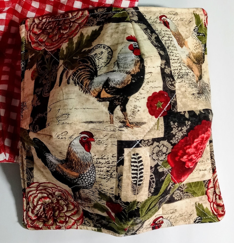 Op de afbeelding: Een gewatteerde stoffen komhoes met een haan- en bloemenontwerp. De stof heeft hanen, rode bloemen en tekst op een cr&egrave;mekleurige achtergrond. Een rood-wit geruite stof is zichtbaar in de linkerbovenhoek.