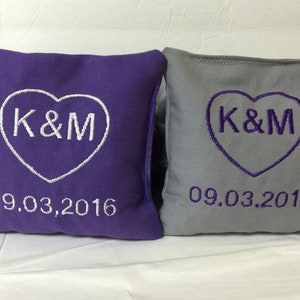 May include: Two square decorative pillows, one purple and one gray. Each pillow features a heart-shaped design with the embroidered initials "K&M" and the date "09.03.2016" in white thread. The pillows are likely for a wedding or anniversary.