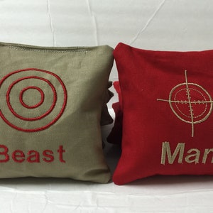 May include: Two square bean bags, one tan and one red. The tan bag has a red embroidered target design and the word "Beast". The red bag has a gold embroidered crosshair target design and the word "Man".
