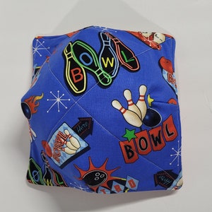 May include: A blue bowl cozy with a bowling theme. The fabric features bowling pins, bowling balls, and the word "BOWL" in a retro font. The cozy is quilted with white thread.