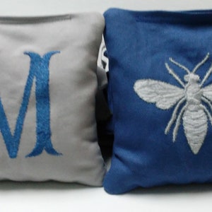 May include: Two square decorative pillows. One is gray with a large blue embroidered letter "M". The other is blue with a white embroidered bee design. Both pillows have a simple, clean design.
