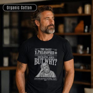 Può includere: Una t-shirt nera a maniche corte in cotone biologico con una stampa grafica bianca e grigia. Il design include il testo The Daily Philosopher, Socrates Asks: But Why?, e Local man questions everything. Again. con un'illustrazione di Socrate.