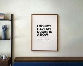 Puede incluir: Un marco con el texto "I DO NOT HAVE MY DUCKS IN A ROW". La impresión tiene un marco marrón y está colgada en una pared color crema. Debajo, un banco de madera oscura con un cojín azul.