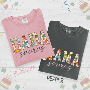 以下が含まれることがあります： 2枚のTシャツ。1枚はブロッサムピンク、もう1枚はペッパーグレーで、恐竜をテーマにしたテキストが特徴です。ピンクのシャツには「DADA saurus」、グレーのシャツには「MAMA saurus」と書かれています。テキストはカラフルな恐竜のイラストで埋められています。