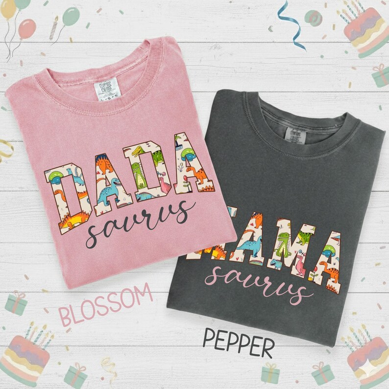 以下が含まれることがあります： 2枚のTシャツ。1枚はブロッサムピンク、もう1枚はペッパーグレーで、恐竜をテーマにしたテキストが特徴です。ピンクのシャツには「DADA saurus」、グレーのシャツには「MAMA saurus」と書かれています。テキストはカラフルな恐竜のイラストで埋められています。