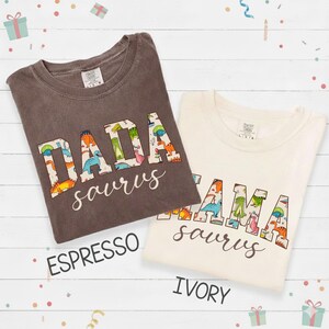 以下が含まれることがあります： エスプレッソ色とアイボリー色の2枚のTシャツが表示されています。エスプレッソのシャツには、恐竜をテーマにした文字で白いスクリプトで「DADA saurus」と書かれています。アイボリーのシャツには、同じ恐竜のデザインで「MAMA saurus」と書かれています。これらはカジュアルウェアのTシャツです。