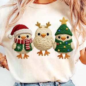 May include: A cream t-shirt with three crocheted chick designs. One chick wears a Santa hat and scarf, another has antlers, and the third is dressed as a Christmas tree. The chicks have orange feet and beaks.