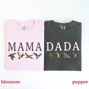 Pu&ograve; includere: Una t-shirt rosa chiaro con la scritta MAMA in grigio scuro e una t-shirt grigio scuro con la scritta DADA in rosa chiaro. Entrambe le magliette presentano una grafica di quattro germani reali in volo sotto il testo. Hanno un girocollo.