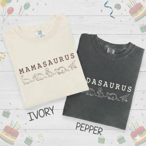 May include: Two t-shirts, ivory and pepper, with dinosaur graphics. The ivory shirt reads "Mamasaurus" and the pepper shirt reads "Dasaurus". Both shirts have a crew neck and short sleeves. The word "Ivory" and "Pepper" are printed below the shirts.
