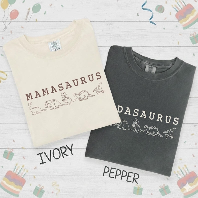 May include: Two t-shirts, ivory and pepper, with dinosaur graphics. The ivory shirt reads "Mamasaurus" and the pepper shirt reads "Dasaurus". Both shirts have a crew neck and short sleeves. The word "Ivory" and "Pepper" are printed below the shirts.