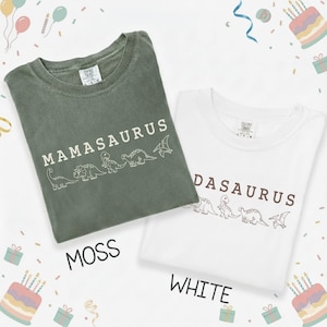 May include: Two t-shirts, one moss green and one white, are displayed. The moss green shirt says "MAMASAURUS" with dinosaur illustrations. The white shirt says "DASASAURUS" with similar dinosaur illustrations. The word "MOSS" and "WHITE" are below the shirts.