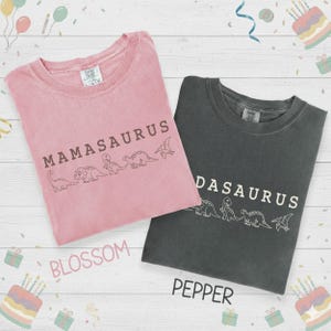 May include: Two t-shirts, one blossom pink and one pepper gray, are displayed. Each shirt has a dinosaur graphic and the words "MAMASAURUS" and "DASAURUS" printed on them. The shirts are on a white wooden surface.