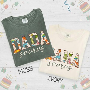 以下が含まれることがあります： モスグリーンとアイボリーの2枚のTシャツが表示されています。モスグリーンのシャツには「DADA saurus」、アイボリーのシャツには「MAMA saurus」と書かれています。テキストは恐竜をテーマにしたフォントです。