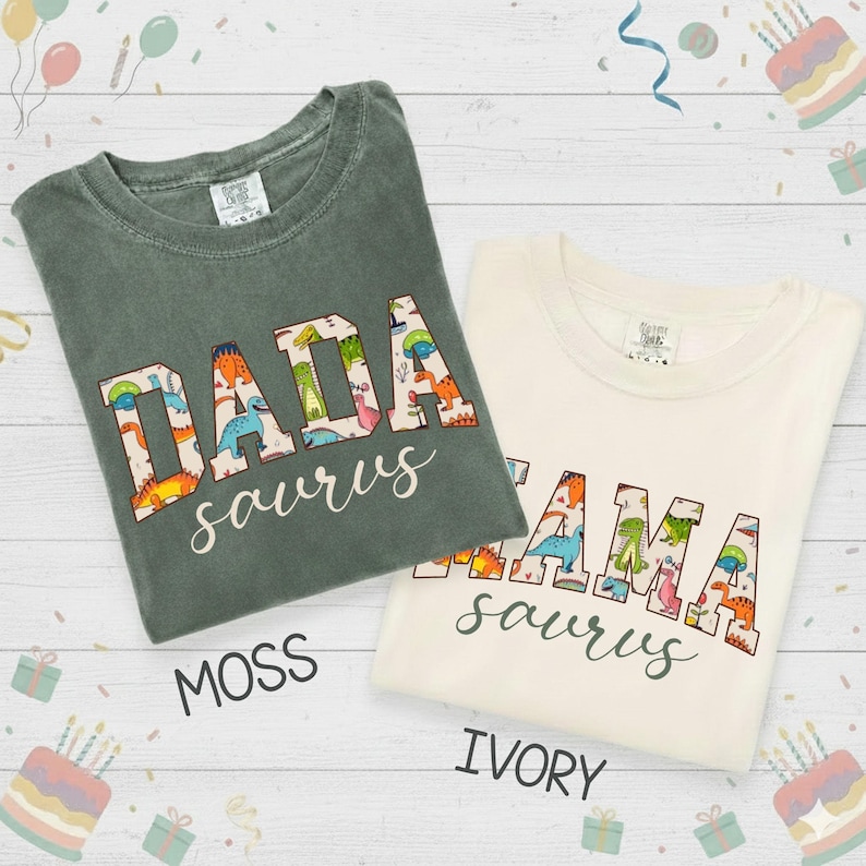 以下が含まれることがあります： モスグリーンとアイボリーの2枚のTシャツが表示されています。モスグリーンのシャツには「DADA saurus」、アイボリーのシャツには「MAMA saurus」と書かれています。テキストは恐竜をテーマにしたフォントです。