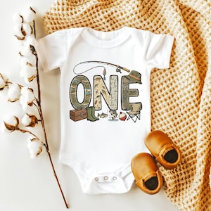 May include: White baby bodysuit with a fishing-themed "ONE" graphic. The design includes a fishing rod, tackle box, boots, and a hat. Brown leather baby shoes are next to the bodysuit. The bodysuit is on a beige knitted blanket.