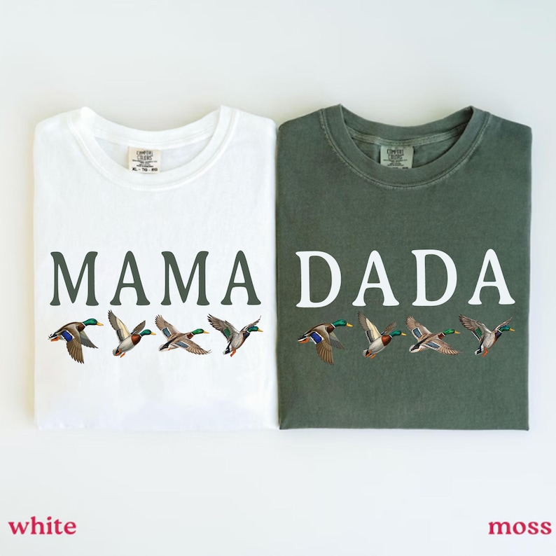 Pu&ograve; includere: Una t-shirt bianca con la scritta MAMA e una t-shirt verde muschio con la scritta DADA. Entrambe le magliette presentano una grafica di quattro germani reali in volo posizionata sotto il testo. Le magliette sono affiancate.