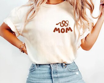 May include: Ivory-colored t-shirt with a brown snake design and the word "MOM" in brown. The shirt has short sleeves and is paired with blue jeans. The word "ivory" is printed at the bottom left corner.