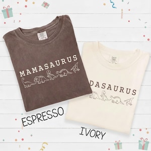 May include: Two t-shirts, one espresso and one ivory, with the word "Mamasaurus" and "Dadasaurus" printed in white. Each shirt features a line of dinosaur illustrations. The shirts are on a white wooden surface.