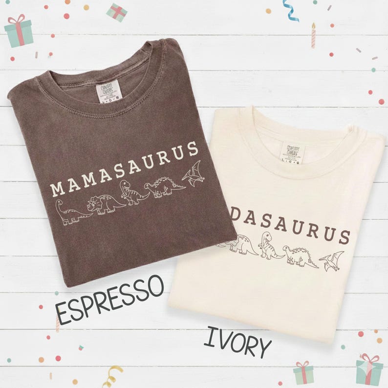 May include: Two t-shirts, one espresso and one ivory, with the word "Mamasaurus" and "Dadasaurus" printed in white. Each shirt features a line of dinosaur illustrations. The shirts are on a white wooden surface.