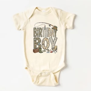 May include: Cream-colored baby bodysuit with a fishing-themed graphic. The words "BIRTHDAY BOY" are prominently displayed, with illustrations of fishing gear, including a rod, hat, and tackle box. A small folding chair and fish complete the design.