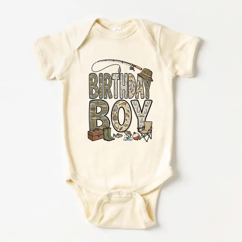 May include: Cream-colored baby bodysuit with a fishing-themed graphic. The words "BIRTHDAY BOY" are prominently displayed, with illustrations of fishing gear, including a rod, hat, and tackle box. A small folding chair and fish complete the design.