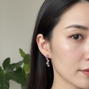 May include: A close-up of a person's ear wearing a pair of dangle earrings. The earrings feature a series of pink and clear gemstones in a vertical arrangement. The gemstones vary in size and shade, creating a gradient effect. The earrings are set in a light-colored metal.