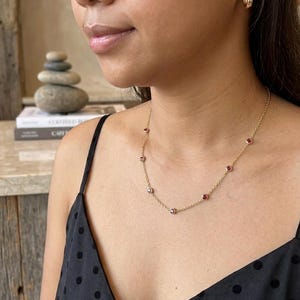 May include: A delicate gold necklace with small, round, red gemstones evenly spaced along the chain. The necklace is worn against a black polka dot top. A pair of gold hoop earrings are also visible.