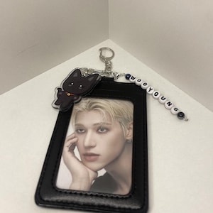 Op de afbeelding: Zwarte leren ID-kaarthouder met een foto van een persoon met blond haar. Een zwarte kattenbedel en een kralenbedel met de tekst "WOOYOUNG" zijn met een zilveren sleutelhanger aan de houder bevestigd.