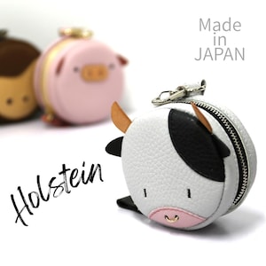 May include: A white and black Holstein cow-shaped coin purse with a silver zip and keyring. The purse has brown horns and a pink nose. The word "Holstein" is written in black cursive. Made in Japan.