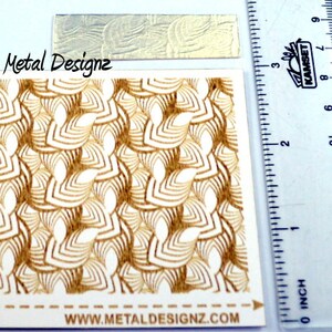 May include: A close-up of a sheet of decorative metal with an Art Deco-style pattern of overlapping scales. The sheet is labeled "ArtDeco-Scales" and "Metal Designz" and includes the website "www.metaldesignz.com".