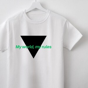 Puede incluir: Camiseta blanca con un triángulo negro y el texto verde "My world, my rules". La camiseta está colgada en una percha blanca contra una pared blanca.