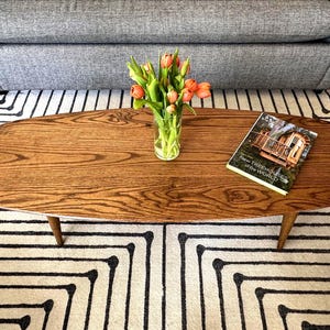 Może przedstawiać: Owalny drewniany stolik kawowy z naturalnym wykończeniem słojów drewna. Szklany wazon z pomarańczowymi tulipanami i książka zatytułowana "New Treehouses of the World" spoczywają na stole. Stół stoi na wzorzystym dywanie.