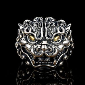 May include: A silver ring depicting a mythical creature's face, with gold-coloured eyes. The ring has detailed carvings and is set against a black backdrop. The design is symmetrical and bold, with a textured surface. The ring's style is ornate and detailed.