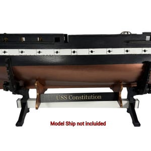 May include: A black and white display stand for a model ship, featuring a horizontal bar with the text "USS Constitution" in gold lettering. The stand has two black vertical supports with adjustable knobs and a white base, holding a ship hull model.