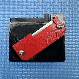 Peut inclure: Un cutter noir avec un boîtier de lame rouge texturé. Le couteau a une lame argentée et est fixé avec des vis argentées. Le couteau est sur une surface bleue.